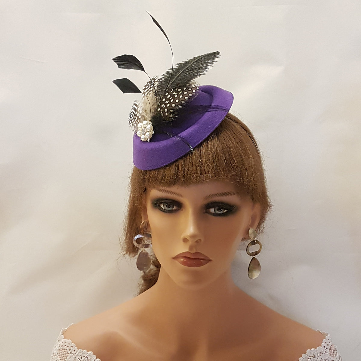 The product in the image is a purple fascinator hat . It features decorative elements including black and white spotted feathers, plain black feathers, and a cluster of faux pearls.