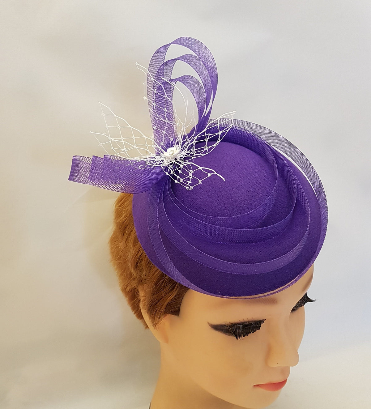 purple felt fascinator hat with a white mesh and ribbon accent. The hat is small and round, and the accent is curled and tied into a bow.