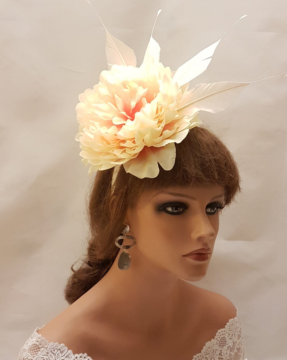 The image features  a floral headpiece or fascinator headband. The headpiece consists of a large, soft-looking flower in a pale peach & cream color,with several delicate petals. The flower is adorned with long slender feathers in a similar light hue.