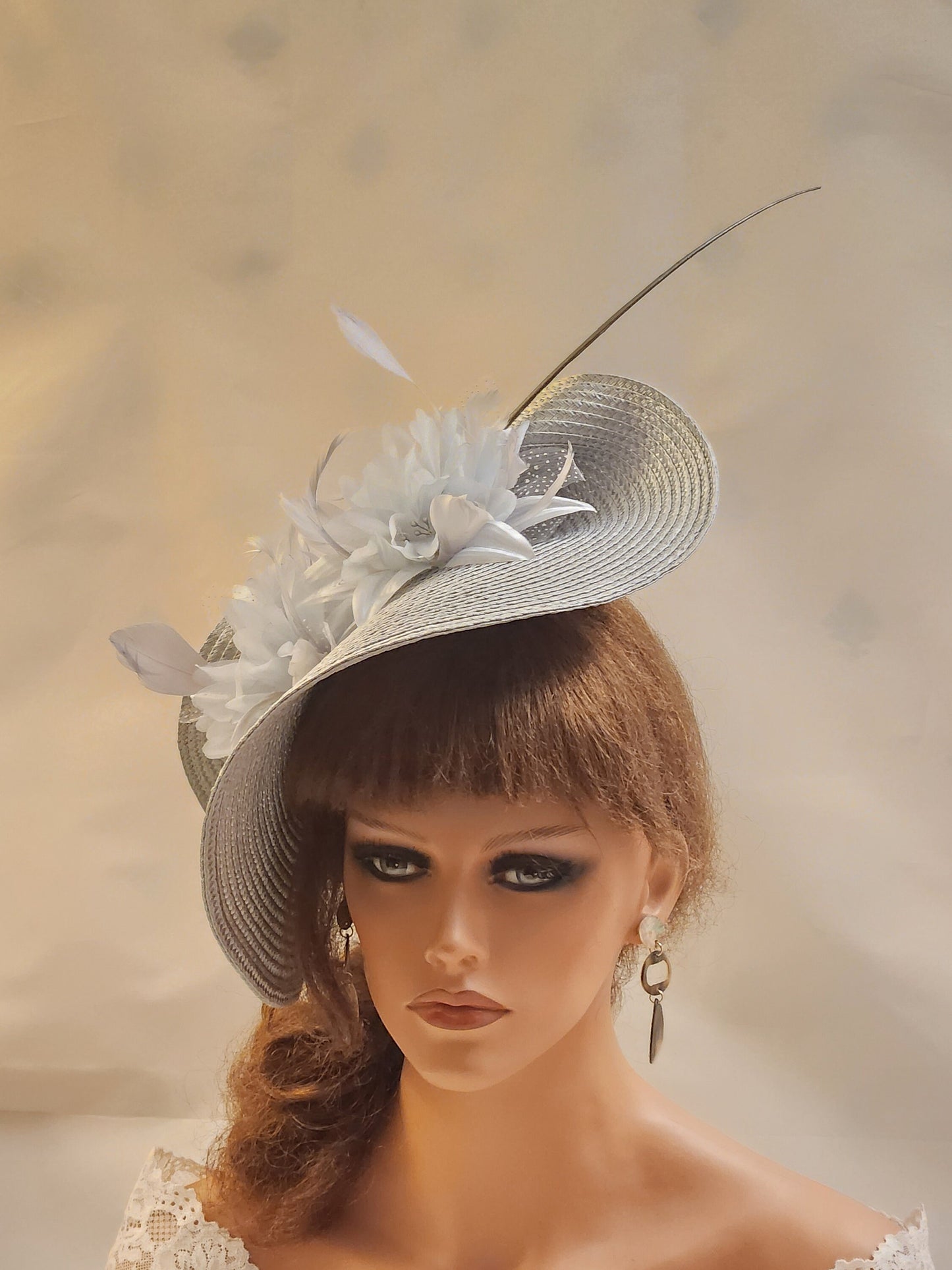A silver straw hat with flowers and long  feathers. The hat is angled to the side and has a long, thin Quill feather .