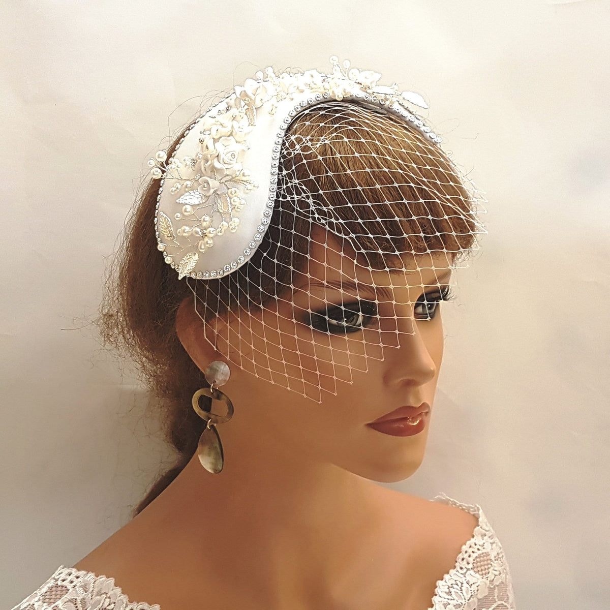 A white satin bridal hat with a pearl and floral embellishment and a white birdcage veil.