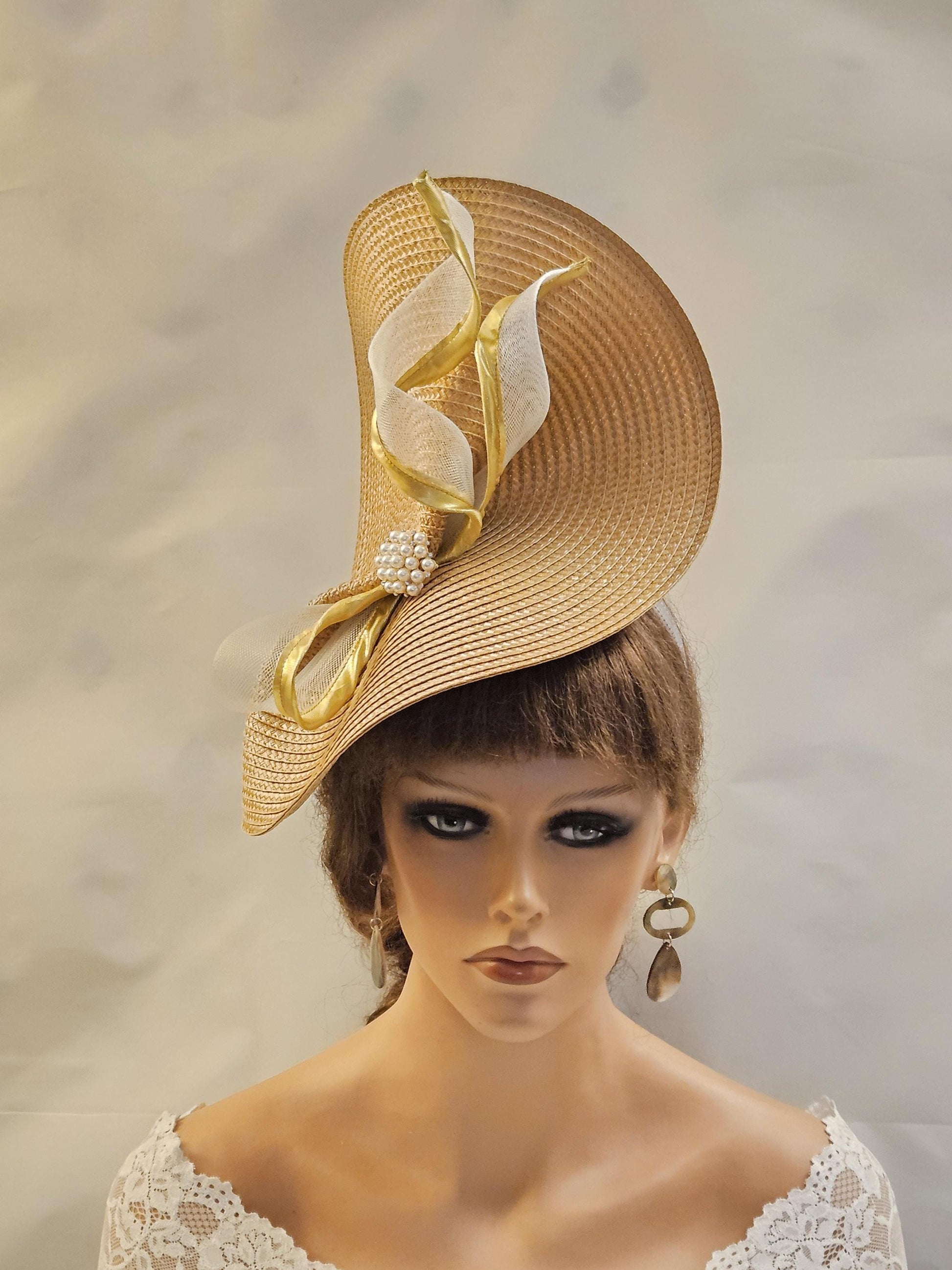 Mother of the bride hat fascinator. Straw weave fascinator with white and gold Crin ribbon accents. The headpiece features a unique, curved design and pearl embellishments. Stylish accessory perfect for weddings or special occasions.