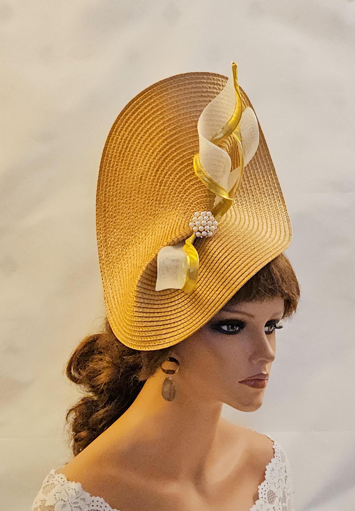 : A gold straw fascinator with off-white and gold ribbon accents. The hat features a unique, sculpted shape and is adorned with pearl-like embellishments.  This elegant headpiece is perfect for weddings, garden parties, or other special occasions.