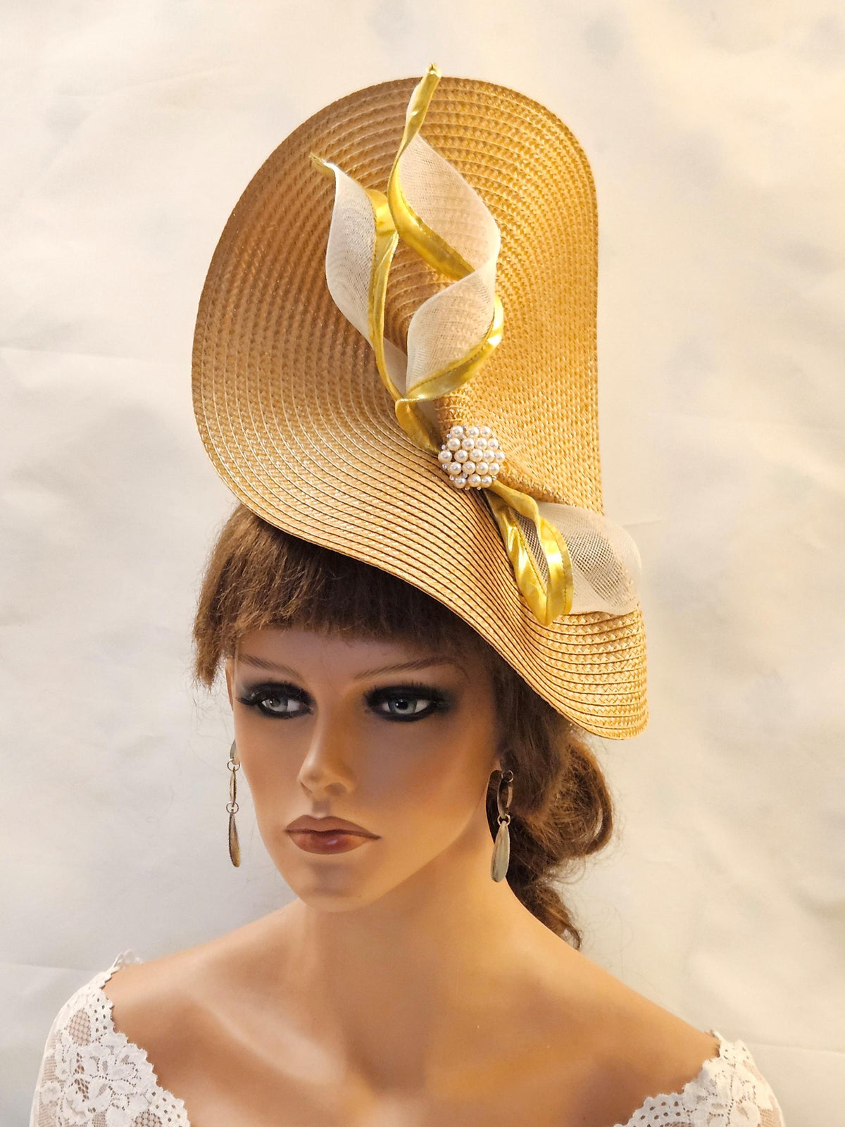 unique sculpted shape hat is adorned with elegant gold and off-white ribbon accents, creating a sophisticated and eye-catching design. A pearl embellishment adds a touch of refined detail. This headpiece is perfect for wedding & special occasions.