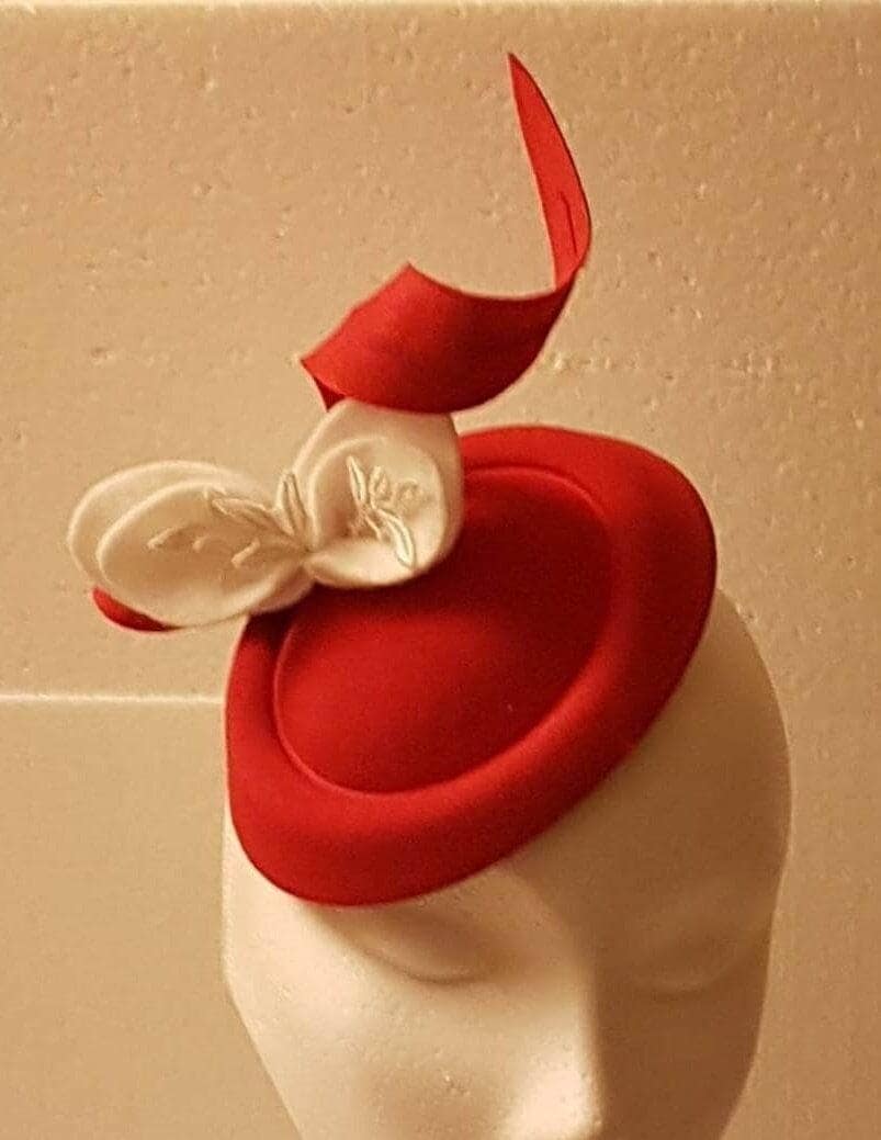 red fascinator .headwear. It features a small, oval base hat that sits atop the head, adorned with a felt embellishment. The decoration includes a sculpted red seril  and a white leaves with subtle embroidery.