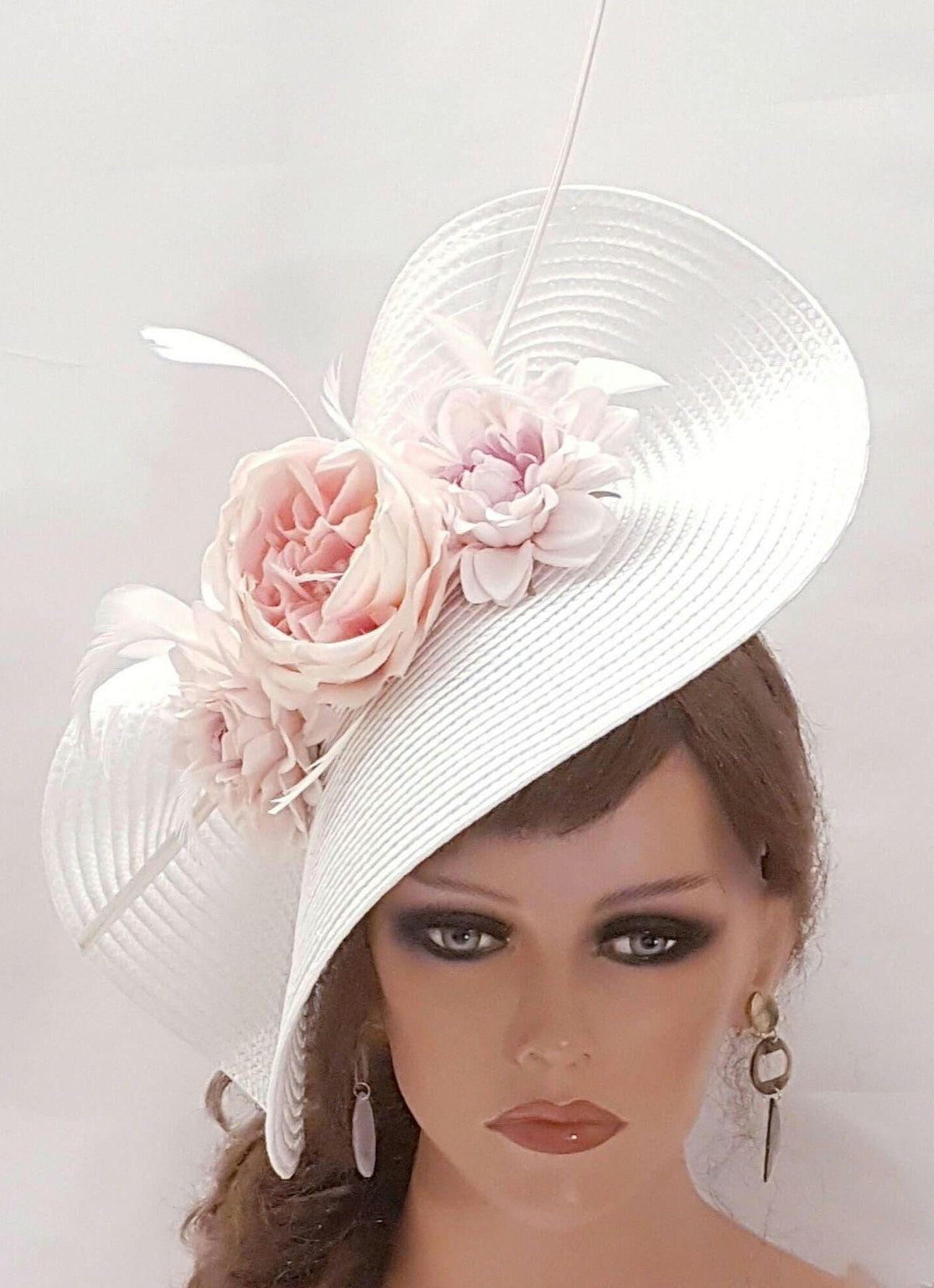 elegant white fascinator hat with an asymmetrical design. It is adorned with large, soft pink  flowers, which are placed prominently on one side. The hat also features delicate feather accents, adding a touch of sophistication.