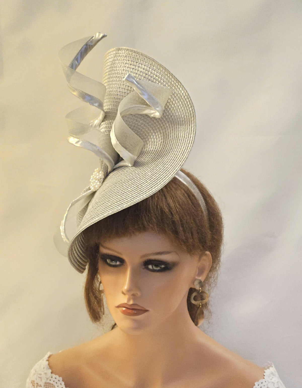 Silver fascinator hat with a unique design. The hat features a large, curved brim adorned with silver ribbon accents creating a swirling,elegant design. Woven straw- headpiece is perfect for weddings, garden parties, special occasions.