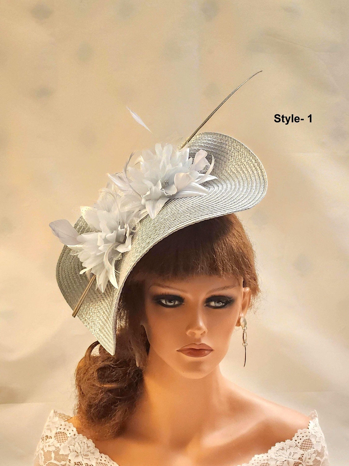 A silver fascinator hat with a white flower and feather design. The hat is angled to the side and has a long Quill feather extending from . .
