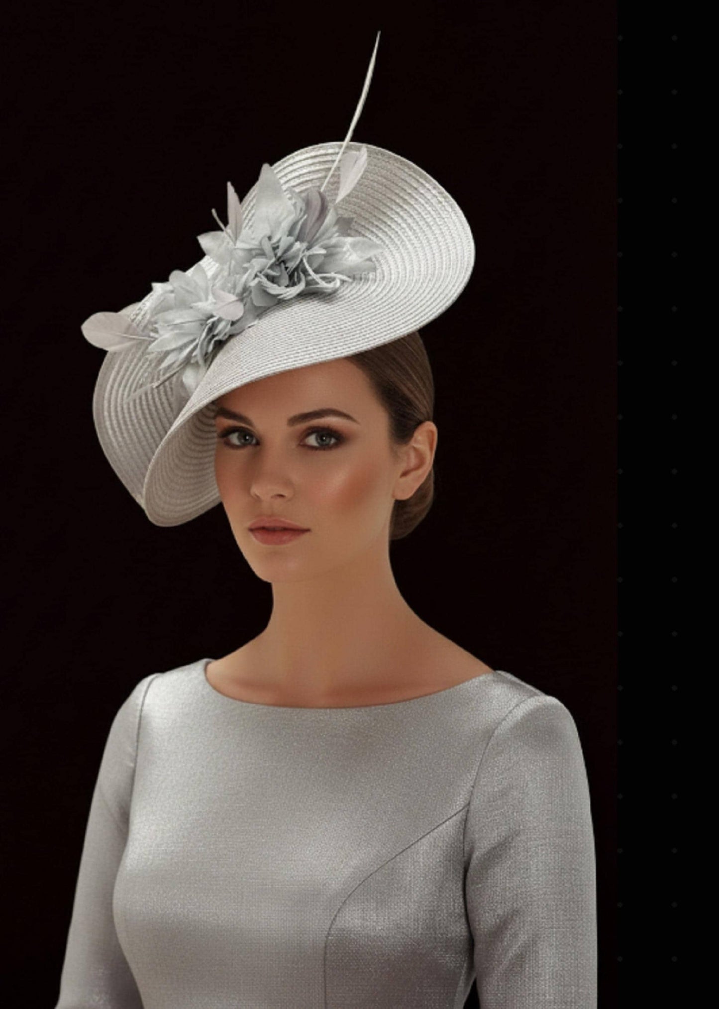 May include: A silver fascinator hat featuring a wide, circular brim with a textured, ribbed surface and a cluster of matching silver fabric flowers and thin, upright feathers attached to the side. The hat is paired with a matching silver long-sleeved top.