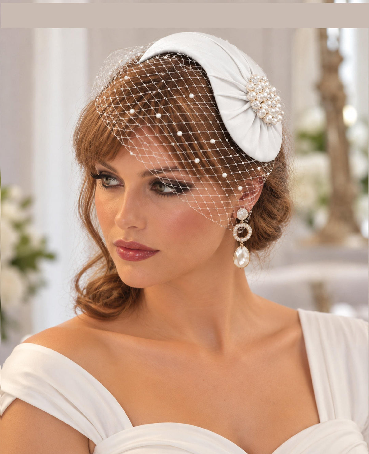 May include: A white fascinator hat featuring a pleated fabric design, a cluster of white pearls at the side, and a white birdcage veil adorned with small white pearls. The headpiece is paired with dangling earrings featuring circular and teardrop shapes.